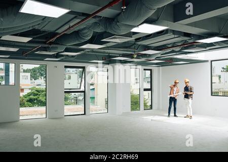 Horizontal extreme long shot of two modern architects standing together in unfinished room discussing building plan, copy space Stock Photo