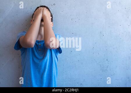 Upset sad boy stand alone and leaning on gray wall. Learning difficulties, family problems, bullying, depression, stress or frustration concept Stock Photo