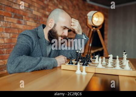 Male chess player playing black figures, thinking process. Chessplayer at board, intellectual tournament indoors. Chessboard on wooden table, strategy Stock Photo