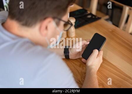 Caucasian man sitting in restaurant and using phone in medical face mask. New Normal concept Stock Photo