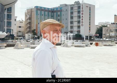 Tel Aviv/Israel-10/10/18: portrait of a smiling old man in a flat cap walking in Tel Aviv on a warm sunny day; Leonardo Beach Hotel on the background Stock Photo