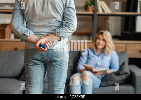 A man holds a box with a ring behind his back and preparing to make a wedding proposal. Love couple in living room together. Happy lifestyle, beautifu Stock Photo