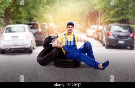 Serviceman in uniform sitting on a pile of tires, street road on background. Repairman, wheel mounting, tyre service Stock Photo