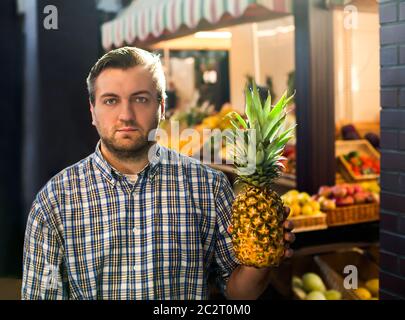 Portrait of smiling man in shirt holds ripe pineapple in hands. Grocery on the background. Stock Photo