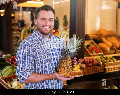 Portrait of smiling man in shirt holds ripe pineapple in hands. Grocery on the background. Stock Photo