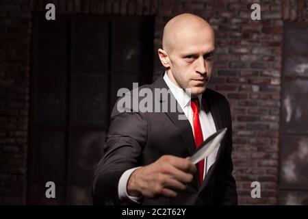 Silent killer, oriental martial arts in action. Bald contract murderer in suit and red tie holds combat knife. Fighting knife is a dangerous weapon in Stock Photo