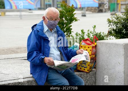 middle aged man, reading newspaper, wearing spotted mask, sat on a wall. Stock Photo