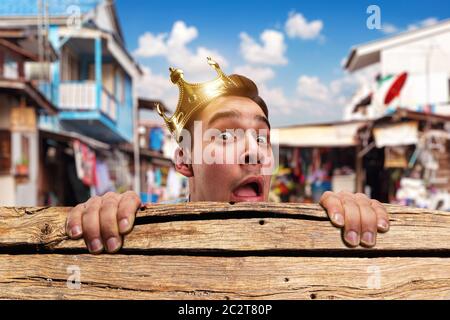 Scared pauper man with crown on the head hidding behind the wooden bench in slums Stock Photo