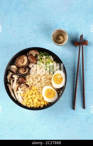 Ramen. Soba noodles with eggs, shiitake mushrooms, and vegetables, overhead shot on a blue background with chopsticks and a cup Stock Photo