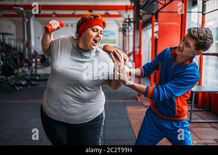 Trainer takes hotdog at fat woman, motivation, hard workout in gym. Calories burning, obese female person in fitness club, fat-burning, sport against Stock Photo