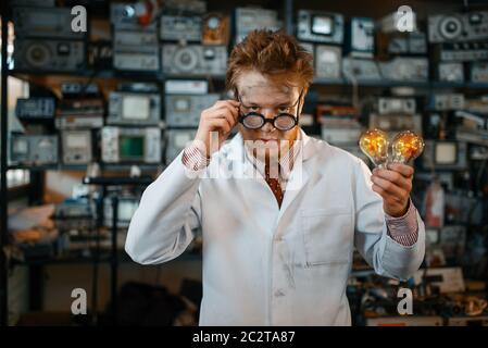 Strange engineer holds electric lamp in laboratory. Electrical testing tools on background. Lab equipment, engineering workshop Stock Photo