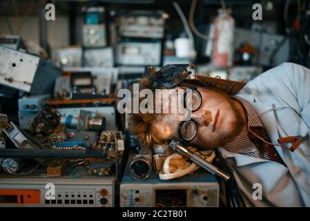 Strange engineer sleeping on devices in laboratory. Electrical testing tools on background. Lab equipment, engineering workshop Stock Photo