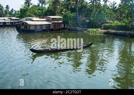 Small houses in a local village located next to Kerala's backwater on a bright sunny day and traditional Houseboat seen sailing through the river Stock Photo