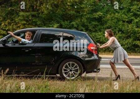 Young woman pushing broken car with man on road, breakdown. Crashed automobile or emergency accident with vehicle, trouble with engine on highway Stock Photo