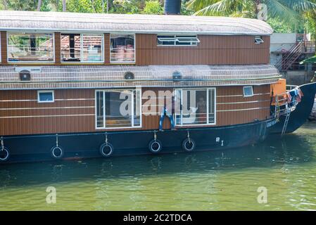 Small houses in a local village located next to Kerala's backwater on a bright sunny day and traditional Houseboat seen sailing through the river Stock Photo