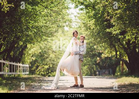 Happy bridegroom raises bride in summer park. man raised and hug woman to her arms. Newly wedded couple in the park. Just marrie Stock Photo