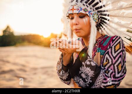 Female American Indian play with sand on sunset. Young girl in headdress made of feathers of wild birds. Traditional costume
