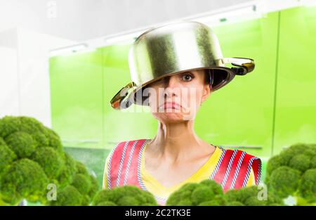 Crazy housewife in apron with a pot on her head among the broccoli, kitchen interior on background. Funny female person cooking Stock Photo