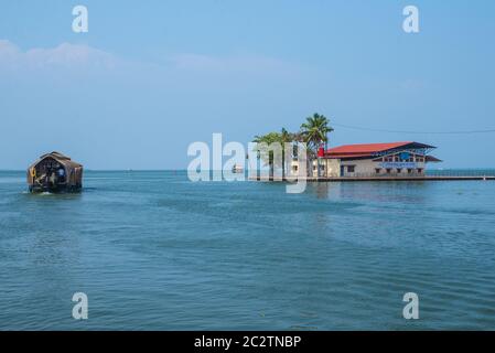 Small houses in a local village located next to Kerala's backwater on a bright sunny day and traditional Houseboat seen sailing through the river Stock Photo