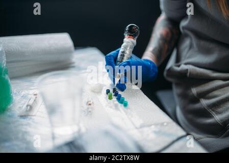 Female tattooist hands in blue sterile gloves holds tattoo machine, professional work tools on background. Tattooing in salon Stock Photo