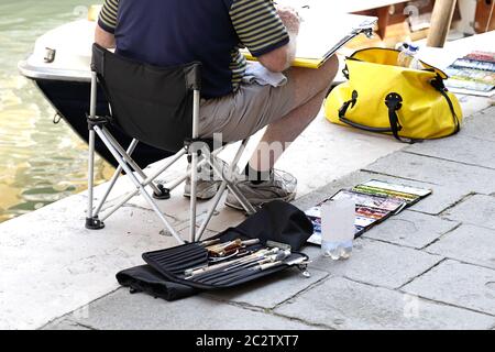 Man artist painting on the street, he is sitting on a chair, tools paint beside his feet Stock Photo