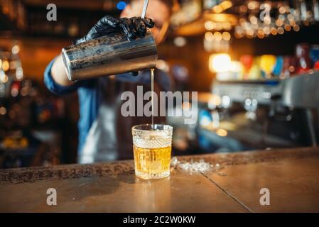 Male bartender in apron pours a drink through a sieve into a glass. Barman at the bar counter. Alcohol beverage preparation Stock Photo