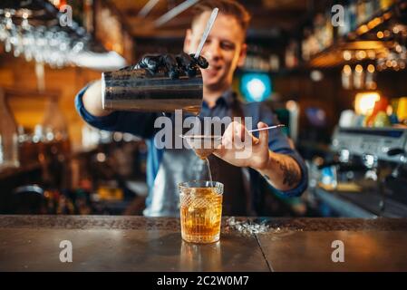 Male bartender in apron pours a drink through a sieve into a glass. Barman at the bar counter. Alcohol beverage preparation Stock Photo