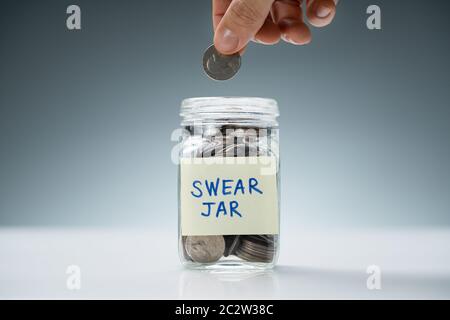 Person's Hand Inserting Coin In Glass Jar With Swear Jar Text Over White Desk Stock Photo