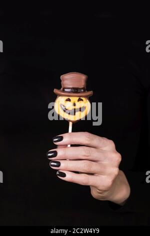 Female hand holding chocolate candy little pumpkin on the stick, black background. Manicure with black nail polish. Copy space Stock Photo