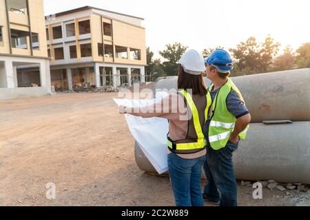 Asian woman construction engineer holding blueprint for checking construction site with technical engineer Building Location on the background, Concep Stock Photo