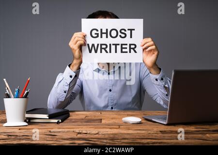 Businessman Hiding His Face Behind Card With Ghost Writer Text Sitting In Front Of Desk Stock Photo
