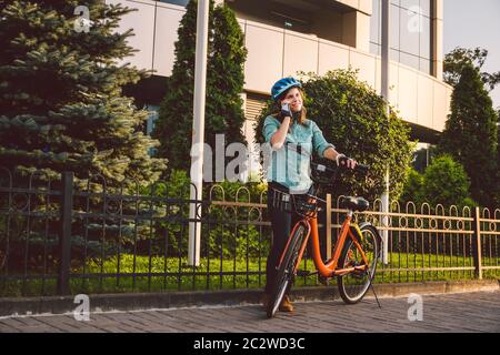 Portrait Of A Cheerful Girl Holding Mobile Phone. Happy smiling student using bike sharing app on smart phone outdoor. City life Stock Photo
