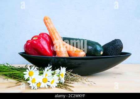 avocado, bell pepper, carrots and cougette in black bowl on wooden table with daisies and grey background Stock Photo