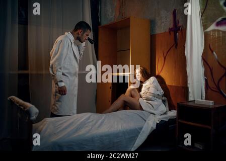 Psychiatrist with torch looks on crazy female patient in strait jacket, mental hospital. Woman in straitjacket undergoing treatment in clinic for the Stock Photo