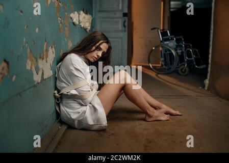 Mad female patient in straitjacket sitting on the floor, mental hospital. Woman in strait jacket undergoing treatment in clinic for the mentally ill, Stock Photo