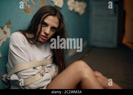 Mad female patient in straitjacket sitting on the floor, mental hospital. Woman in strait jacket undergoing treatment in clinic for the mentally ill, Stock Photo