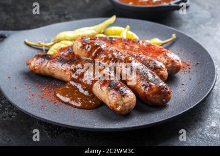 Traditional barbecue German Bratwurst with hot sauce and peperoni as closeup on a modern design cast iron plate Traditional barbecue German Bratwurst with hot sauce and peperoni as closeup on a modern design cast iron plate