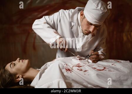 Crazy psychiatrist maniac makes an incision to female patient, experiment in mental hospital basement, bloody walls on background. Victim of mad docto Stock Photo