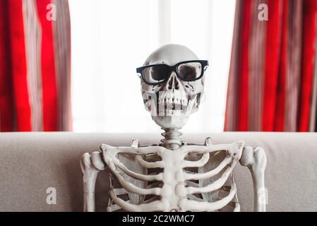 Smiling skeleton in glasses is sitting on couch, window and red curtains on background. Funny joke Stock Photo