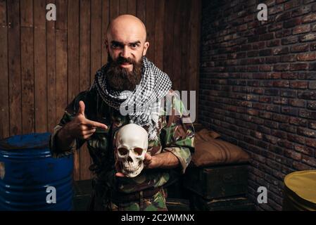 Terrorist in uniform holds human skull in hands, male mojahed. Terrorism and terror horror, bearded soldier in khaki camouflage, barrels of fuel or ch Stock Photo