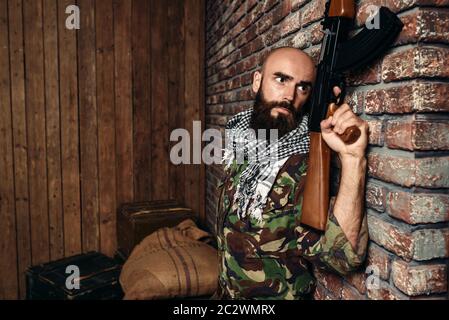 Terrorist in uniform with kalashnikov rifle hiding behind a wall, male mojahed with weapon. Terrorism and terror, soldier in khaki camouflage, boxes o Stock Photo