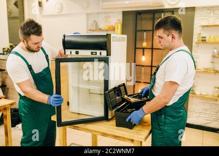 Two workers in uniform repair refrigerator at home. Repairing of fridge occupation, professional service Stock Photo