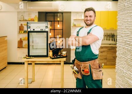 Worker in uniform against refrigerator on the table at home. Repairing of fridge occupation, professional service Stock Photo