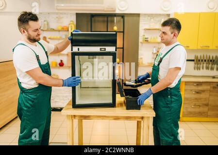 Two workers in uniform repair refrigerator at home. Repairing of fridge occupation, professional service Stock Photo