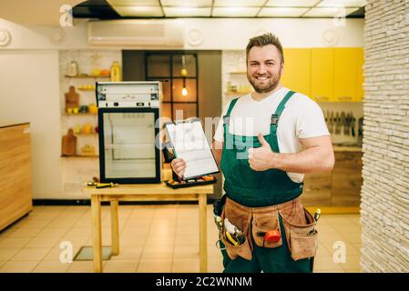 Worker in uniform holds checklist against refrigerator at home. Repairing of fridge occupation, professional service Stock Photo