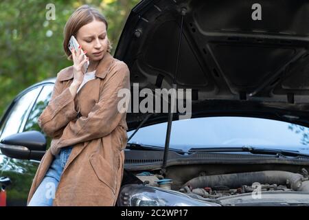 Helpless woman driver calling for help/assistance looking at broken down car, stopped at the roadside. Doesn't understand what happened. Stock Photo