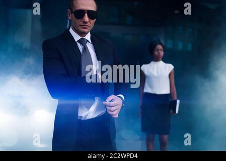 Serious bodyguard in suit, sunglasses and earpiece, female client on background. Security guard is a risky profession, professional guarding of busine Stock Photo