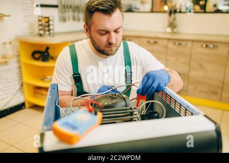 Worker in uniform testing problem with refrigerator at home. Repairing of fridge occupation, professional service Stock Photo