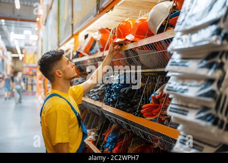 Male builder choosing helmet at the shelf in hardware store. Constructor in uniform look at the goods in diy shop Stock Photo