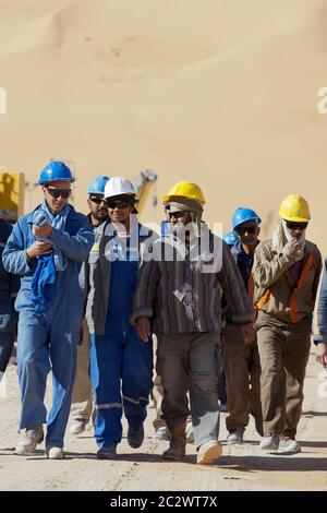 Workers breaking for lunch while working on the construction of a major oil facility in the Sahara desert. A large sand dune rises behind them. Stock Photo